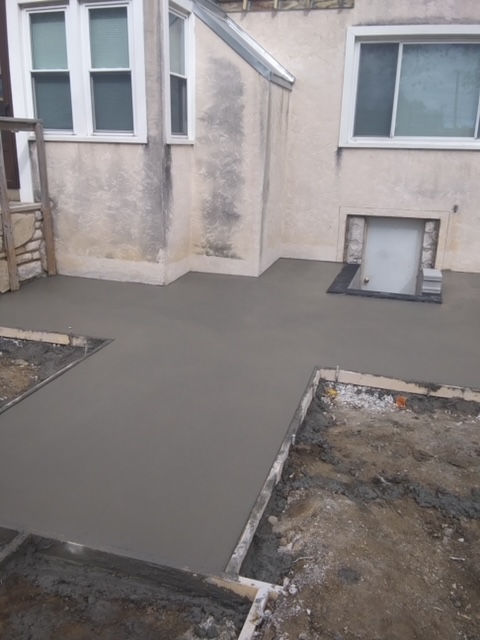 Freshly poured concrete patio extending from the back of a house, framed by wooden forms and patches of bare dirt.