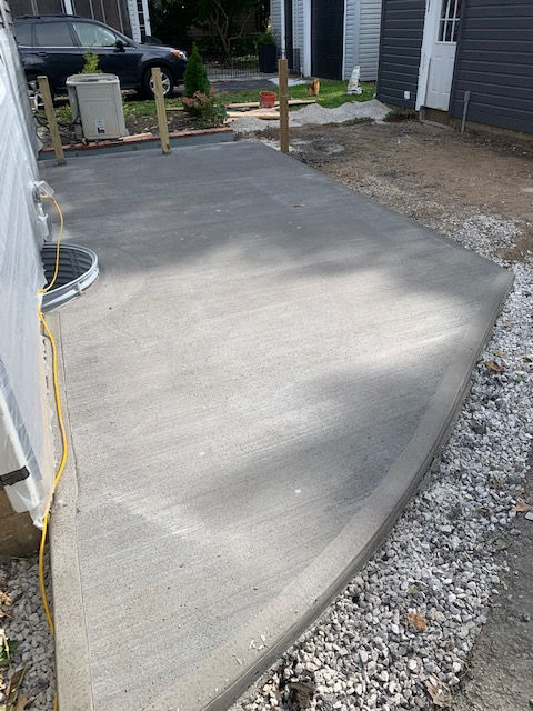 Freshly poured concrete patio next to a house with gravel surrounding the edges and wooden stakes in the background.