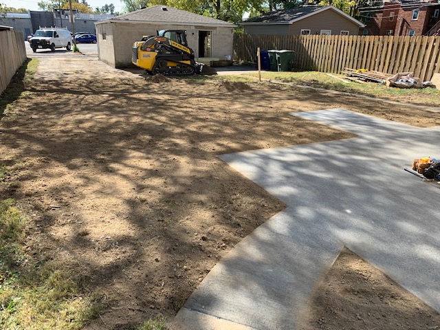 A construction site with a yellow skid steer parked near a concrete slab in a residential backyard.