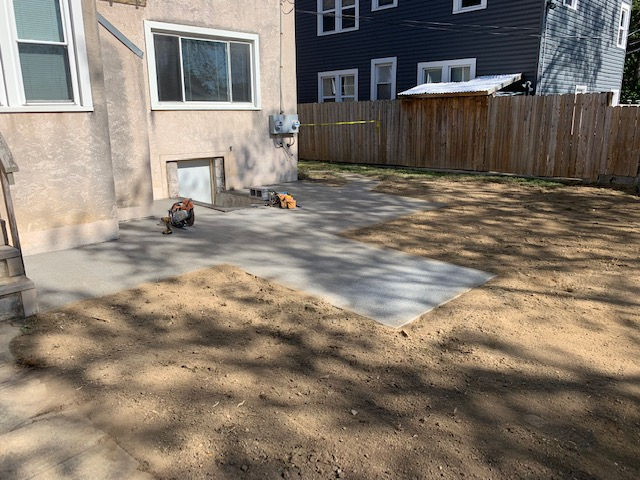 A light-colored house exterior with a newly poured concrete patio in a yard with dirt and shadows from nearby trees.