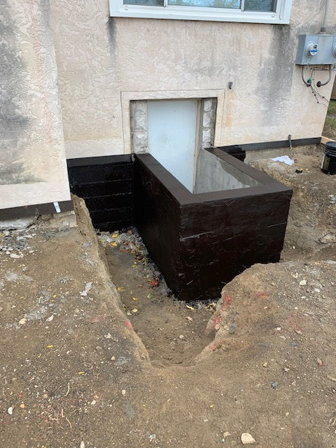 A new dark-coated window well installed against a house foundation with a partially backfilled trench in the dirt yard.