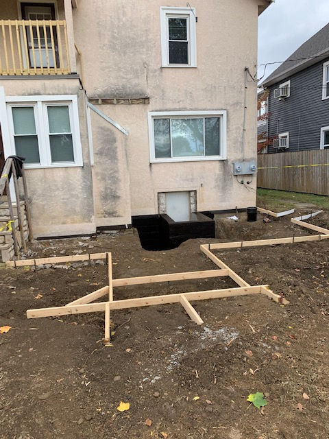 Wooden concrete formwork for a patio being prepared in the dirt-filled backyard of a beige house.