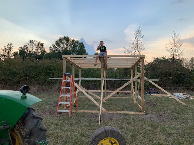 A person sits atop the wooden frame of an uncompleted outdoor structure in a grassy field near a tractor.