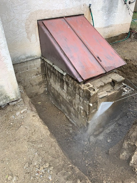 A metal cellar door attached to a house, with a partially excavated concrete block stairwell area in front of it.