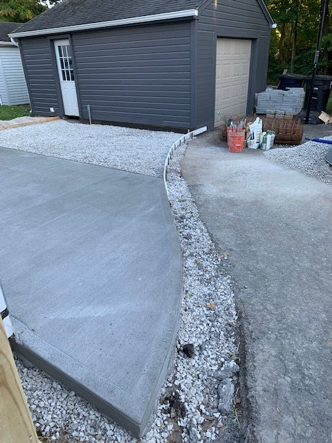 Freshly poured gray concrete patio abutting a gravel path and a dark gray garage in a residential backyard.