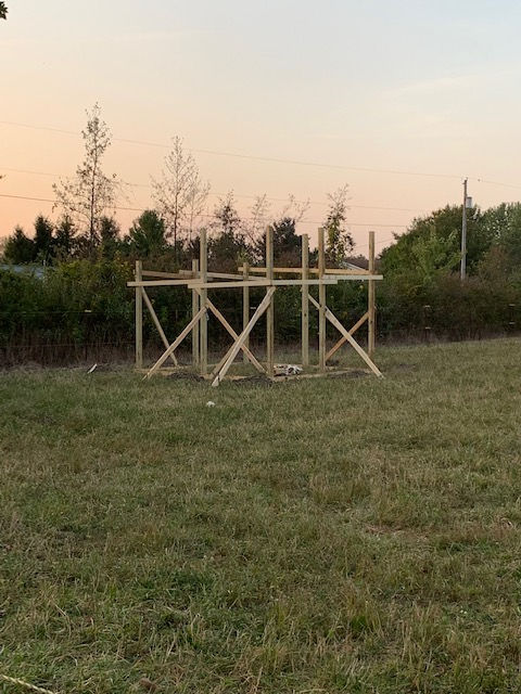 A wooden frame for a small structure stands in an open grassy field during sunset.