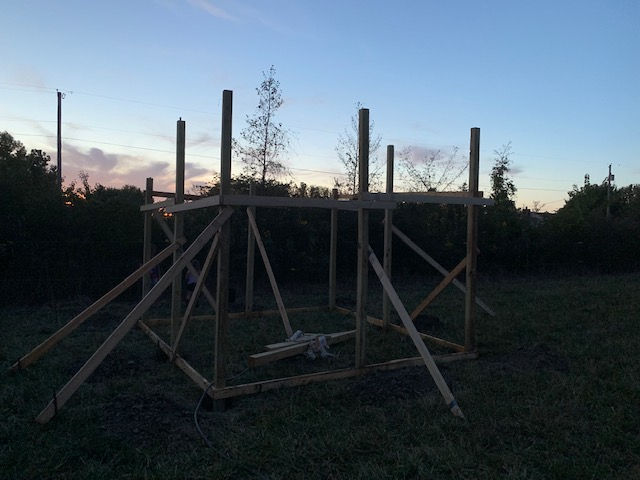 A wooden frame structure stands in a grassy field against a twilight sky, supported by diagonal bracing beams.