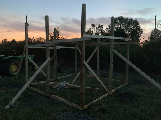 Wooden framing for a small structure standing in a grassy field at sunset, with a tractor visible in the background.