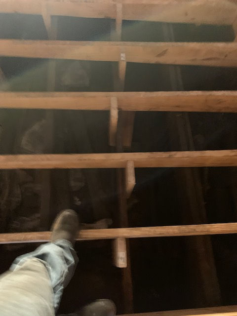 A person’s feet and legs stand on wooden attic joists looking down into a dark crawl space.