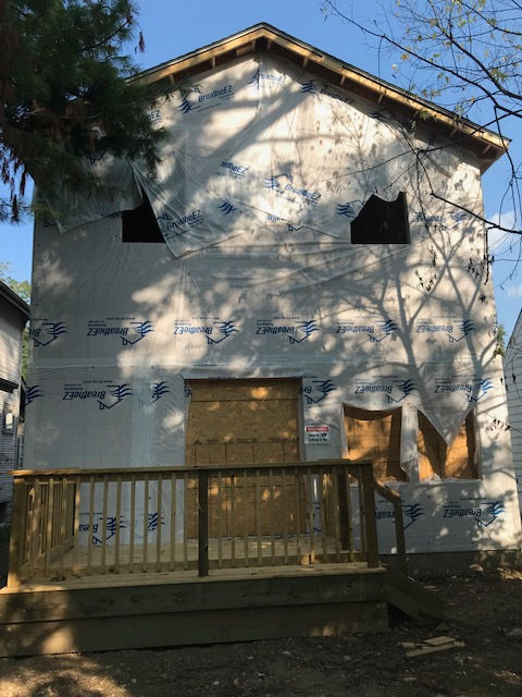 A two-story house under construction, covered in white building wrap with boarded-up windows and a wooden front deck.