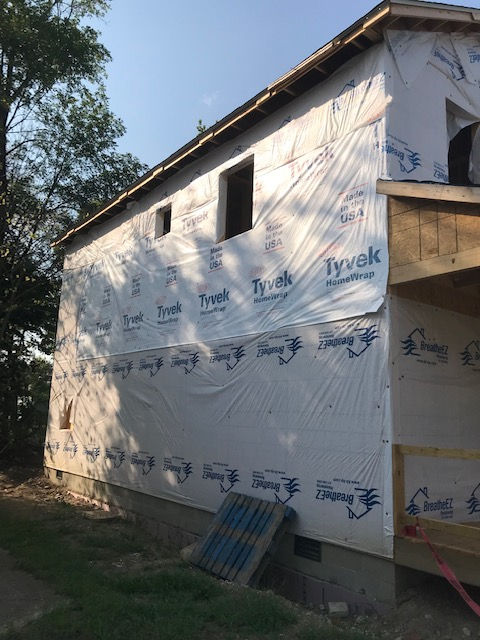 Side view of a two-story house under construction, wrapped in white Tyvek building paper with two open window frames.