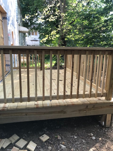 A newly constructed wooden deck featuring vertical railings, viewed from the ground against a backdrop of green trees.