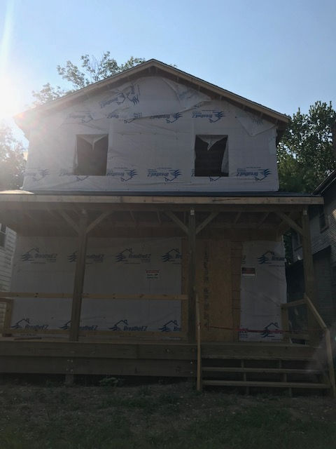 A two-story house under construction, featuring a front porch and exterior walls wrapped in protective white weather barrier.