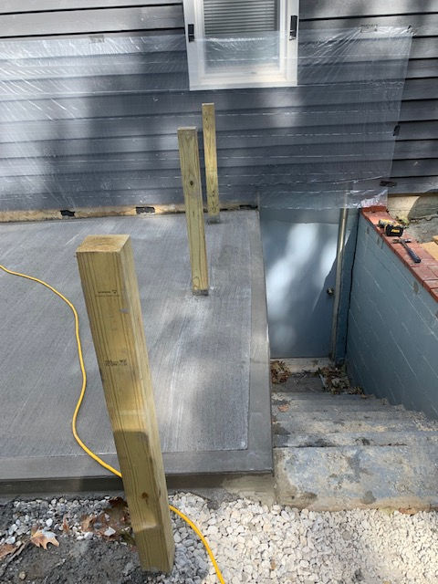 Three wooden posts stand on a grey deck next to a basement stairwell alongside a house with grey siding.