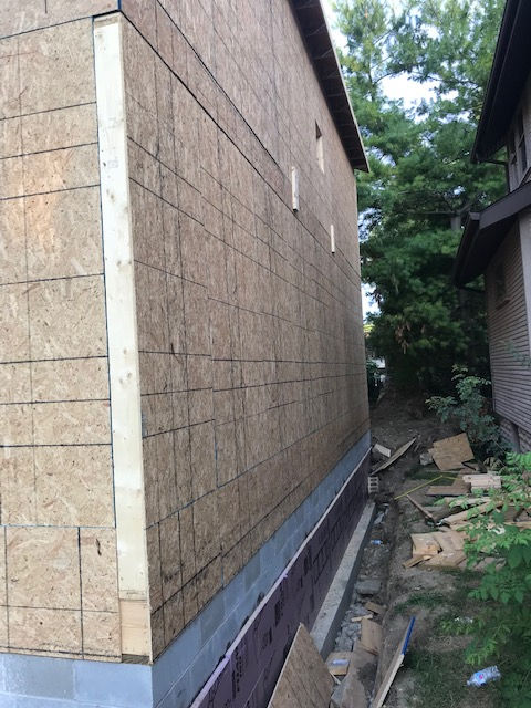 Exterior view of a residential building under construction, featuring plywood walls and a concrete block foundation.