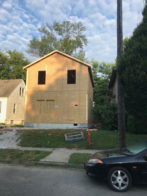A two-story wood-framed house under construction stands on a grassy lot next to a street with a parked black car.