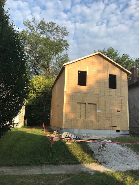 A two-story house under construction with light-colored wooden siding, visible windows, and a concrete foundation.