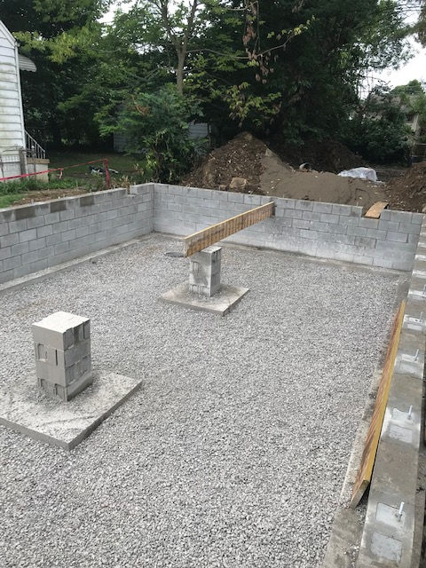 Construction site showing a concrete block foundation and two central support pillars on a gravel base.