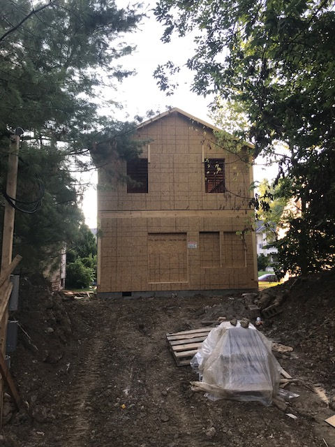 A two-story house under construction, framed with plywood, surrounded by trees and dirt ground.