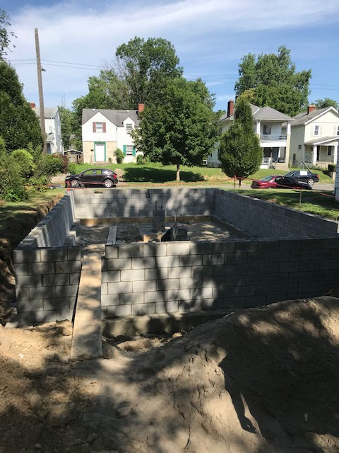 A rectangular foundation made of grey concrete blocks sits in a dirt-filled construction site in front of houses.