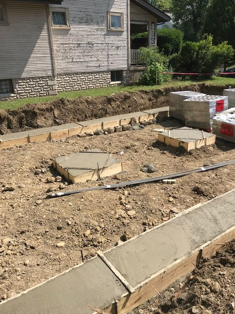Fresh concrete footings and a foundation perimeter wall in a construction trench next to a house.