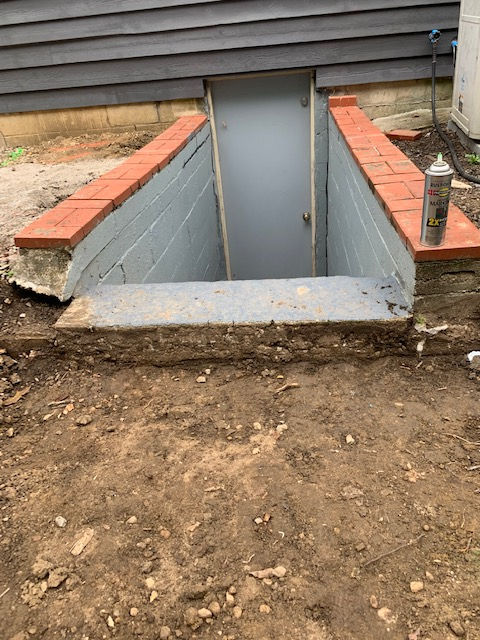 A gray basement stairwell entrance leading to a door, framed by concrete block walls with red brick caps.