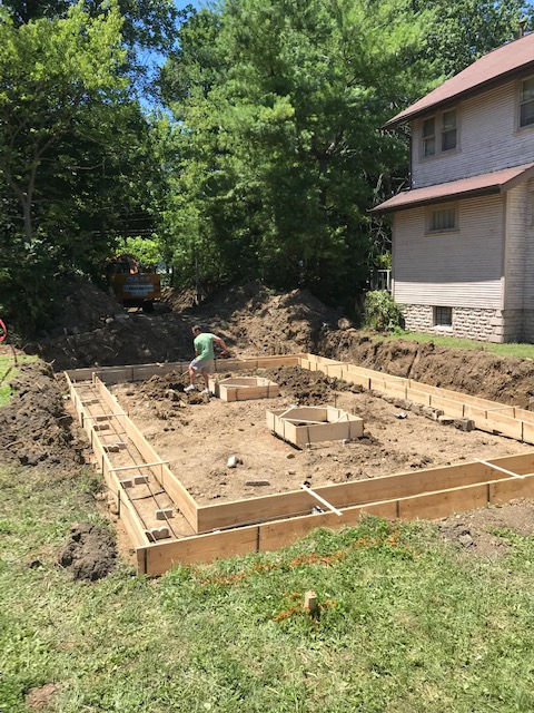 Construction workers set up wooden concrete forms and rebar for a foundation in a residential backyard next to a house.
