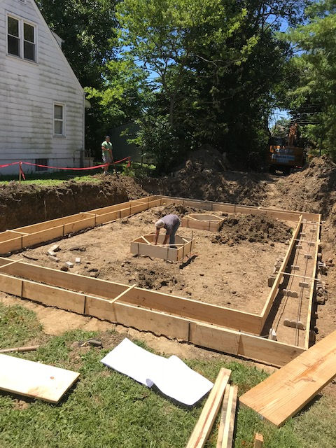 A person works inside wooden foundation forms set in a dug-out area next to a white house.