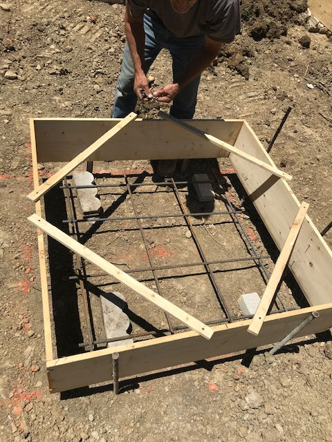 A person prepares a wooden form and rebar grid on a dirt construction site for a concrete footing.