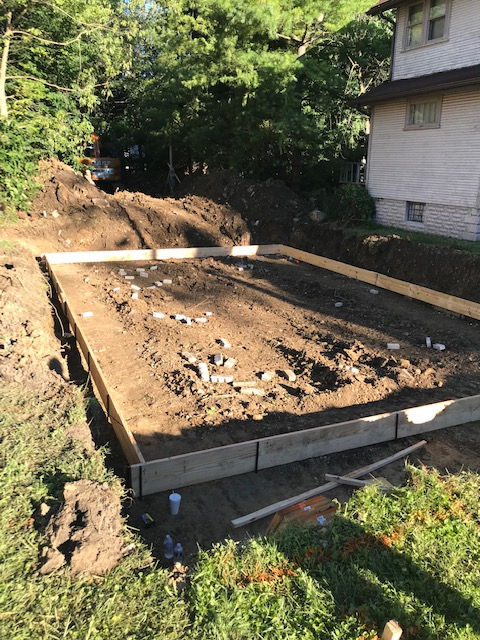 A rectangular concrete foundation frame sits in an excavated yard next to a two-story house, with loose soil inside.