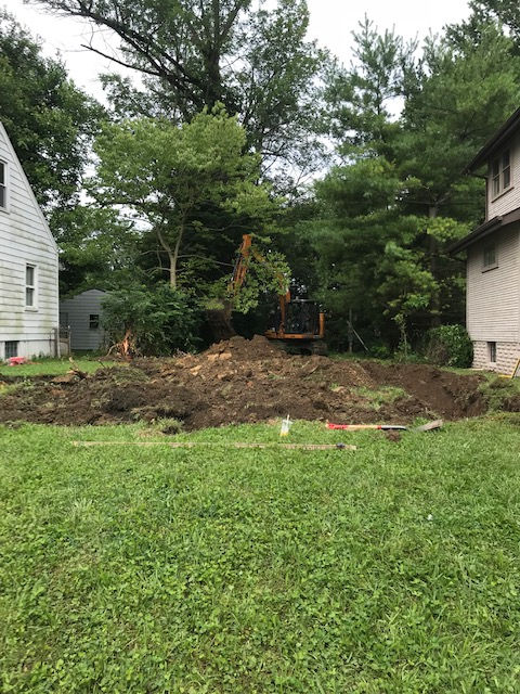 An excavator digs into the ground in a grassy residential backyard between two houses surrounded by trees.