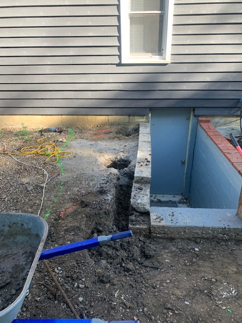A construction site featuring an excavated trench along the exterior foundation of a gray-sided house near a basement door.