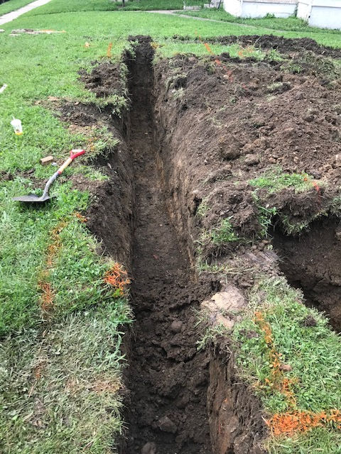 A long, deep trench dug into a grassy lawn, with orange marking paint and a shovel on the side.