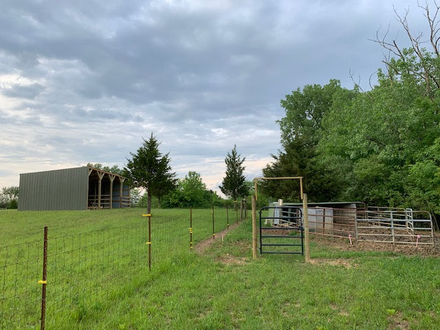 A metal pole barn and a gated enclosure sit in a grassy pasture under a cloudy sky.