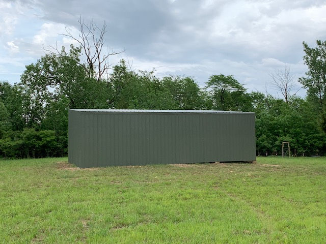 A simple, dark green metal storage building sits in an open, grassy field bordered by trees under a cloudy sky.