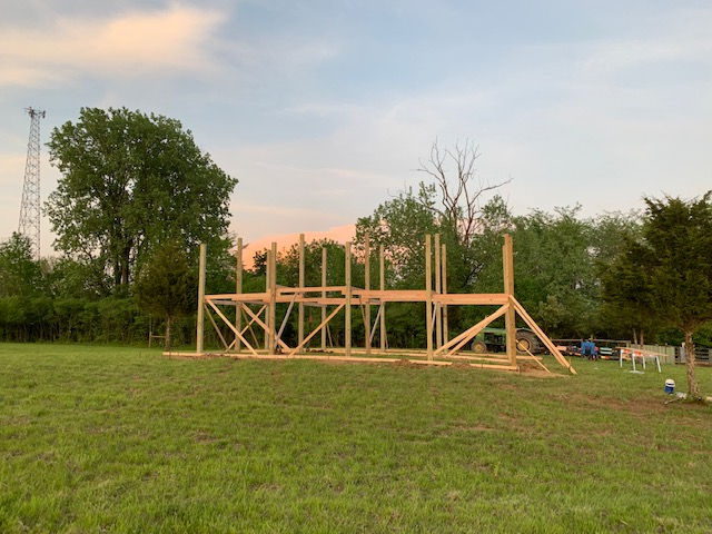A wooden post-and-beam frame structure stands in a grassy field under a sunset sky.