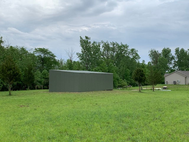 A rectangular, olive-green metal storage building sits on a grassy field with trees and a house in the background.