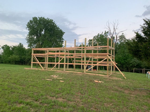 A wooden post-and-beam frame for a building stands in a grassy field under a cloudy sky.