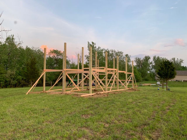 A wooden frame structure under construction stands in a grassy field surrounded by trees at sunset.