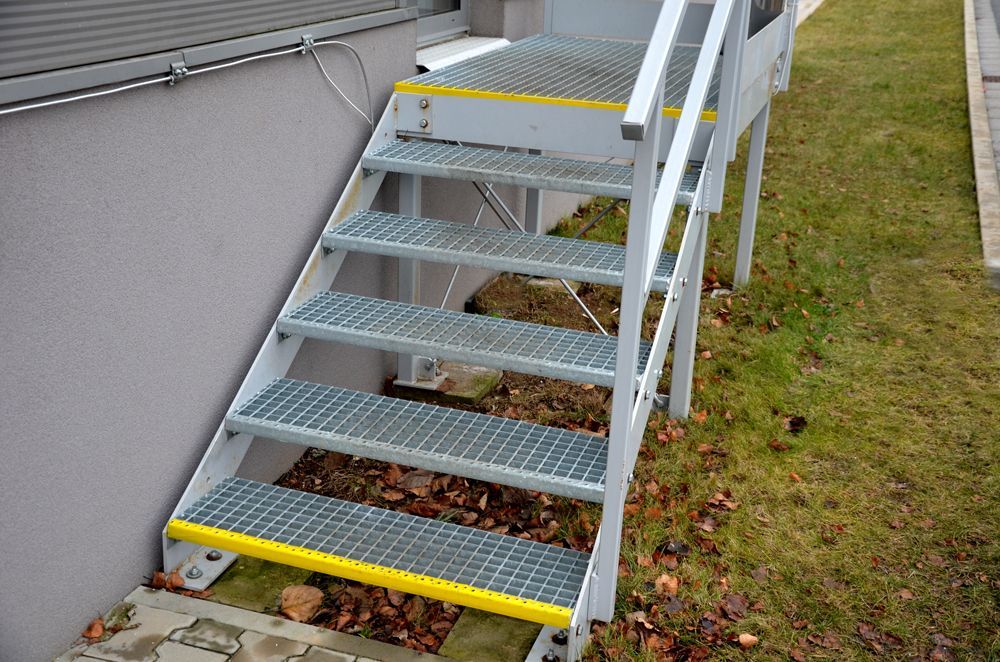 Metal staircase with open-grate steps and yellow safety edges, installed outdoors against a grey building wall.