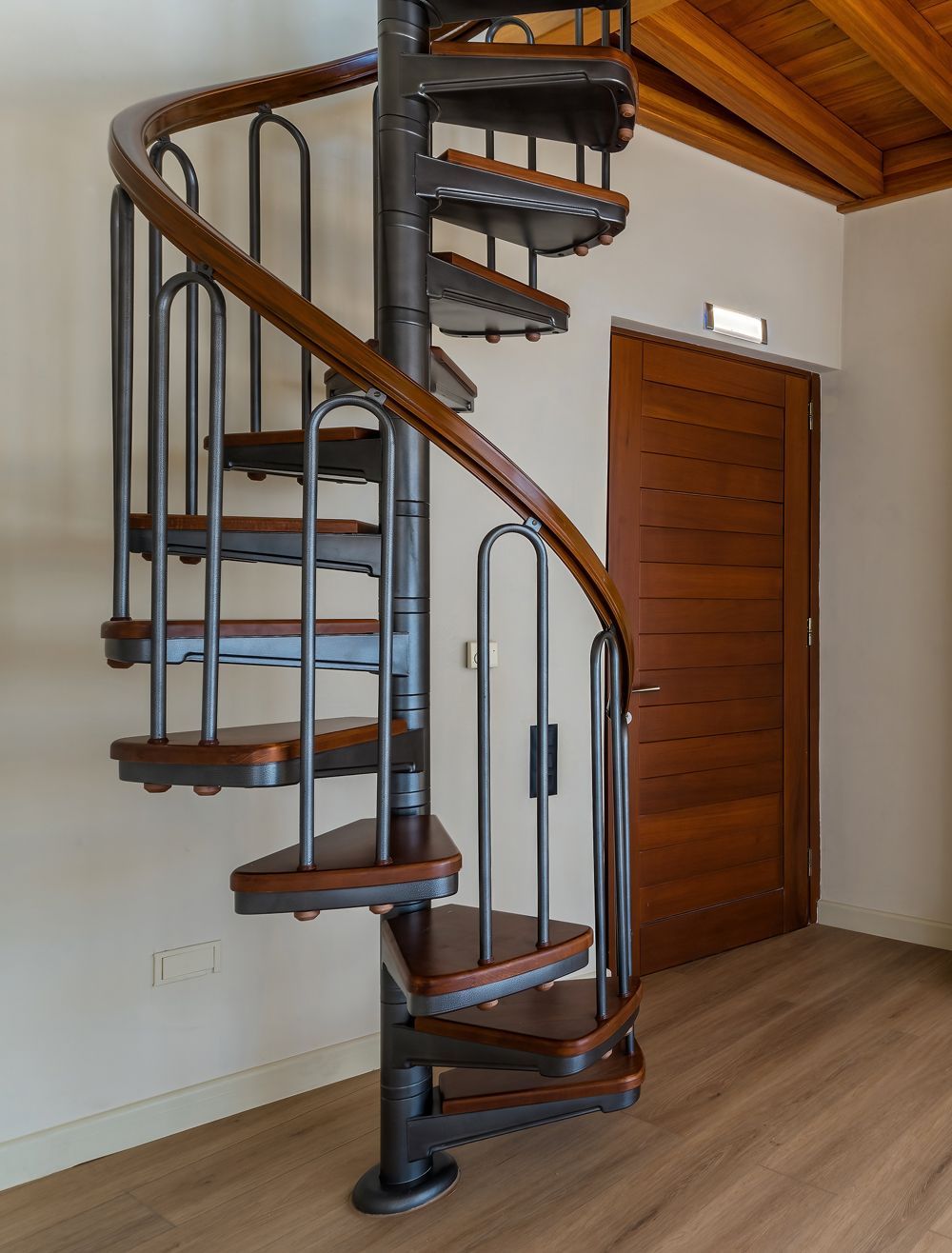 A spiral staircase with a central metal pillar, wooden treads, and a curved wooden handrail in a room with a wooden door.