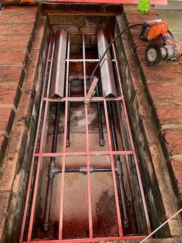 A metal gas burner installation inside a brick-lined trench, with a handheld grinder resting on the side.