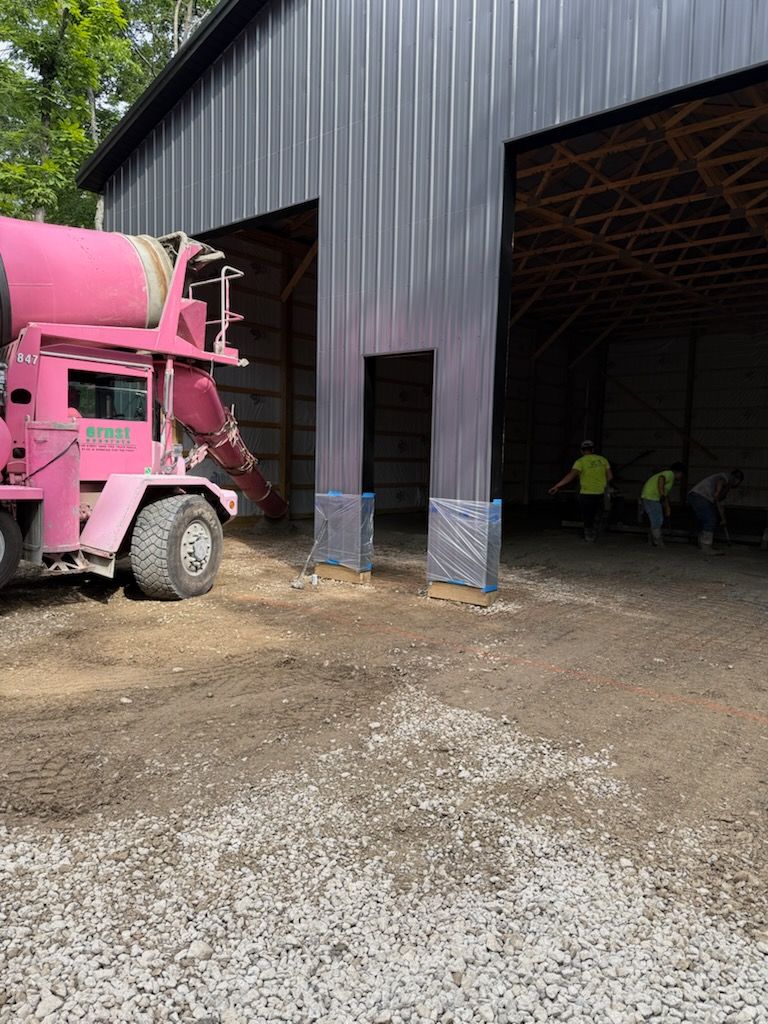 Pink cement mixer truck beside a dark metal barn with open bays and workers inside