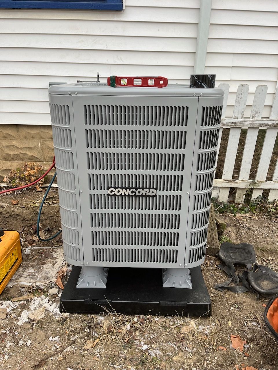 A gray Concord air conditioner unit sitting on a black base outdoors, topped with a red spirit level.