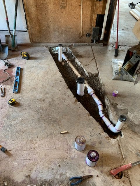 A trench in a concrete floor shows newly installed white PVC plumbing pipes, surrounded by construction tools and supplies.