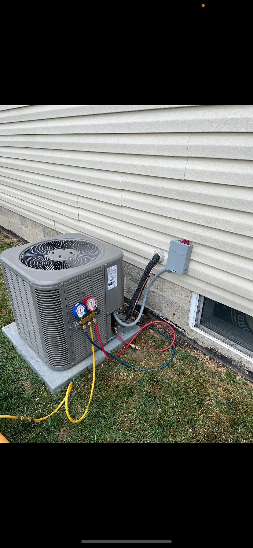 An outdoor air conditioning unit sits against a beige vinyl-sided wall, connected by hoses to a manifold gauge set.