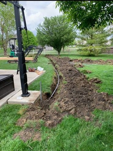 A trench dug through a green lawn, with a pipe inside and a small excavator parked nearby on a concrete patio.