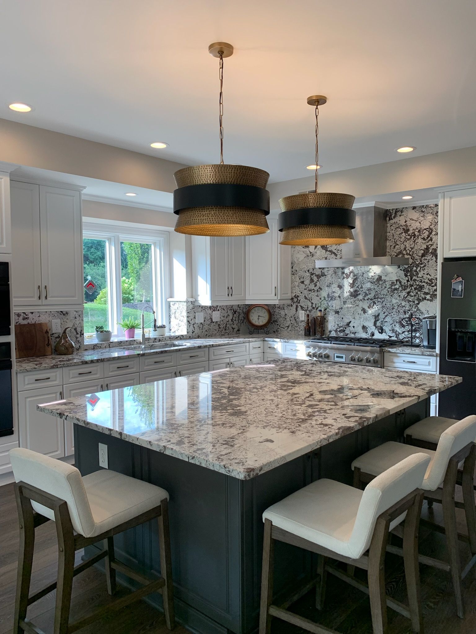 A kitchen island with stools sits in front of white cabinets, granite countertops, and two hanging drum pendant lights.
