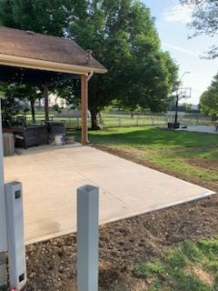 A backyard scene with a covered concrete patio, a grass lawn, large trees, and a basketball hoop in the distance.