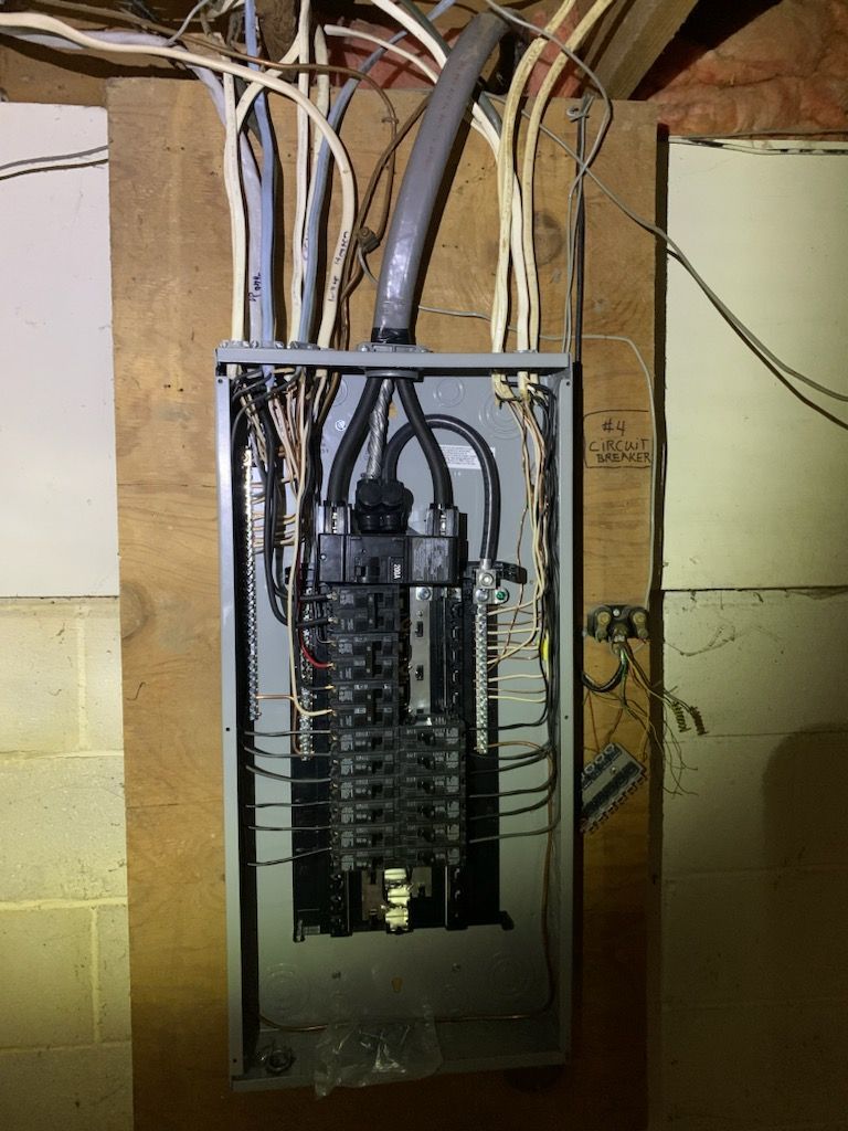 An open residential electrical breaker panel mounted on a wooden board against a cinder block wall.